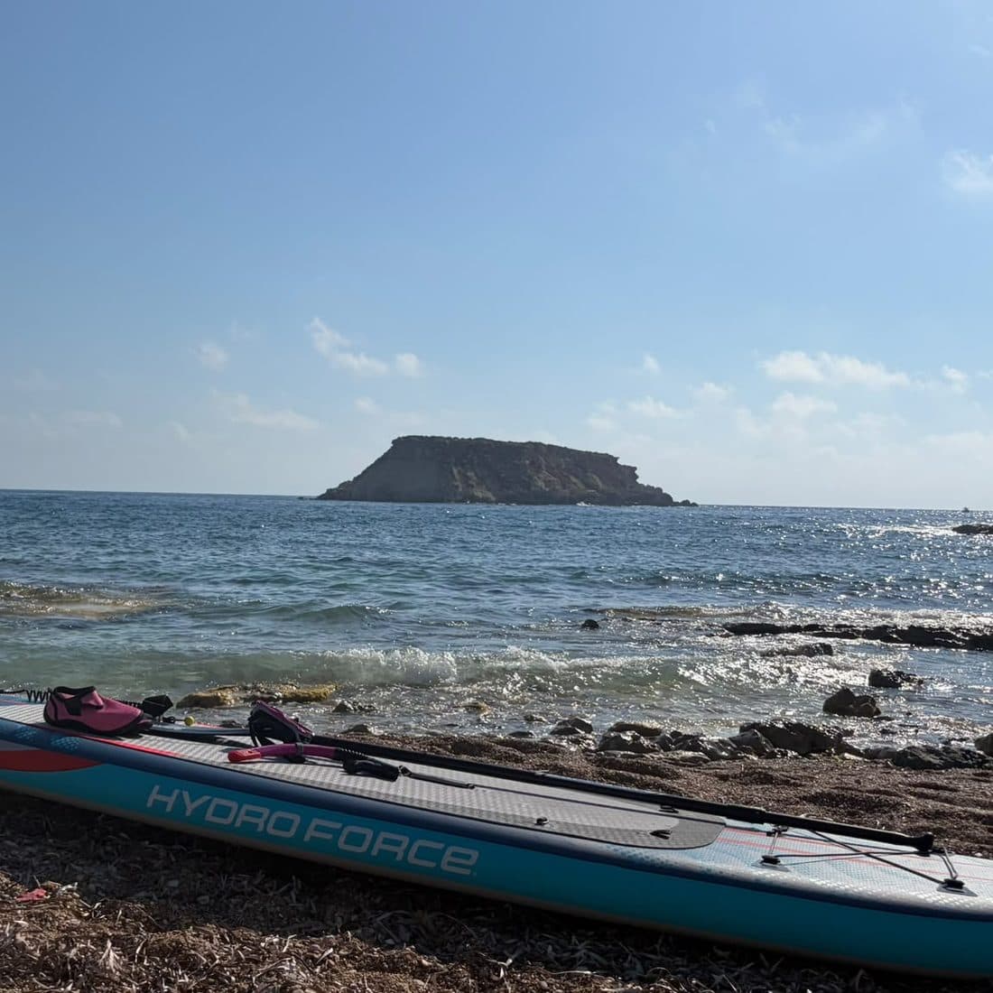 A paddleboard on a pebble beach, facing a small rocky island in a blue sea