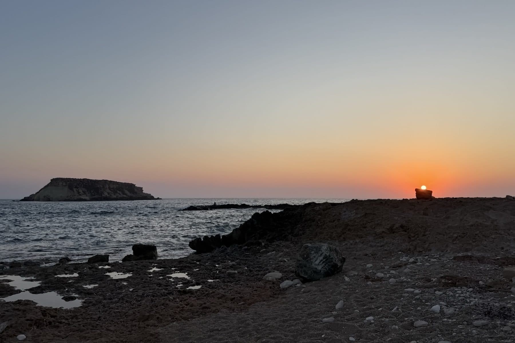 The sun setting over the sea behind a rocky shore, with a small flat island offshore